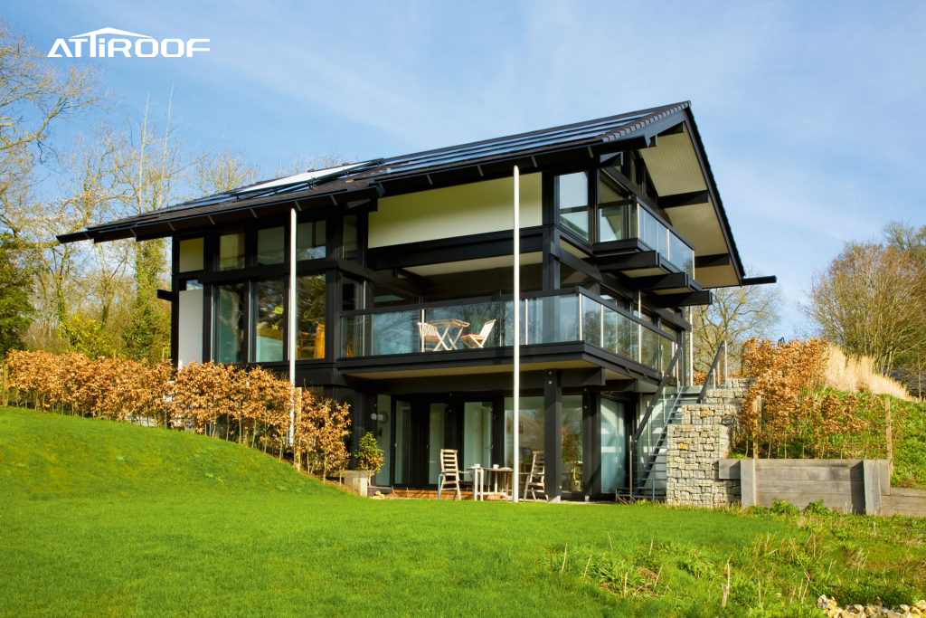 Una casa moderna y ecológica de nueva construcción con grandes ventanales de vidrio y un elegante techo de tejas de resina sintética negra, frente a un paisaje natural.