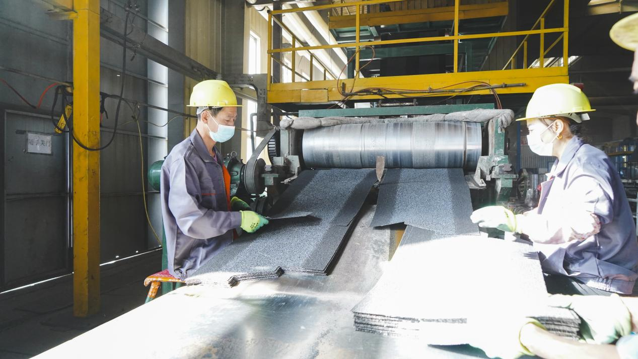 Dos trabajadores, vestidos con monos y cascos, trabajan intensamente en la maquinaria dentro de la fábrica, manipulando un montón de tejas de asfalto negras.